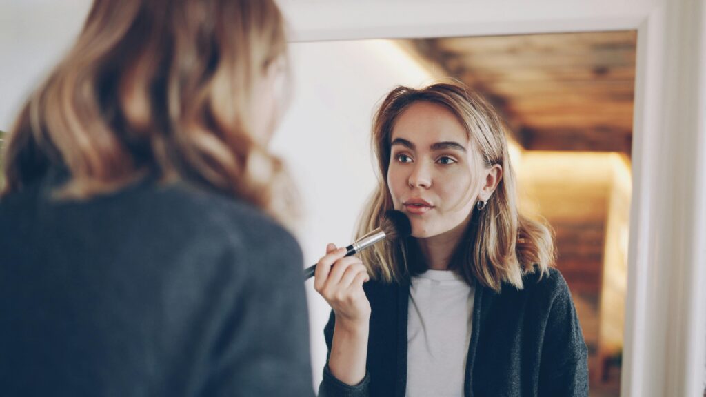 Young woman applying makeup with a brush while looking in the mirror indoors.