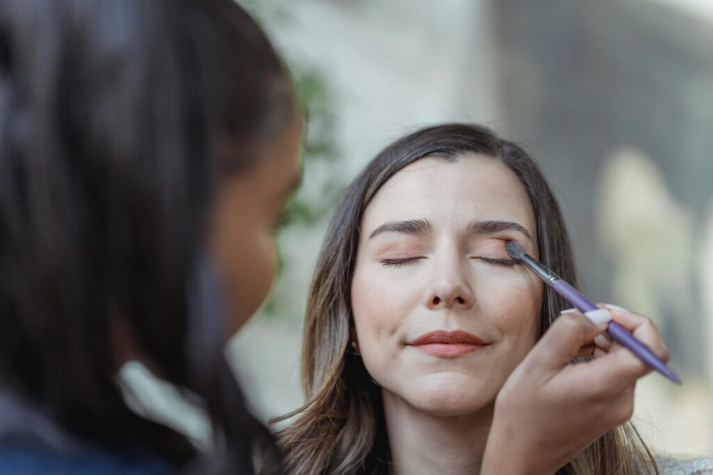 Crop young ethnic female makeup artist using brush while applying eyeshadow on eyelid of client with closed eyes in light beauty salon