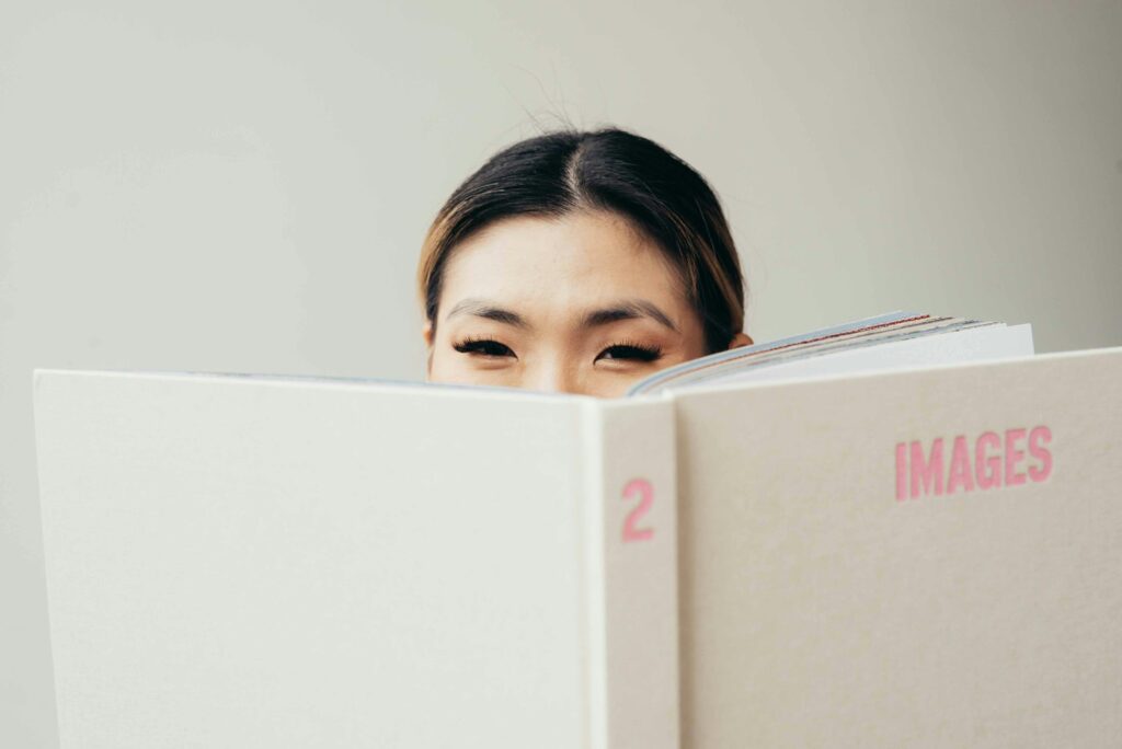 Pretty Asian woman holding opened white book with pink title while looking at camera in studio on white background